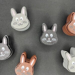 May include: Six gray, brown, and white bunny-shaped figurines with pink ears and white eyes. The figurines are made of a soft, textured material.