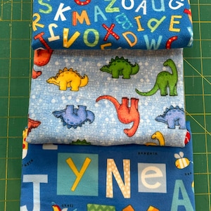 May include: Three folded fabric pieces with colorful patterns. The top piece is blue with a repeating alphabet design. The middle piece is light blue with dinosaur illustrations. The bottom piece is blue with letters and animal illustrations.