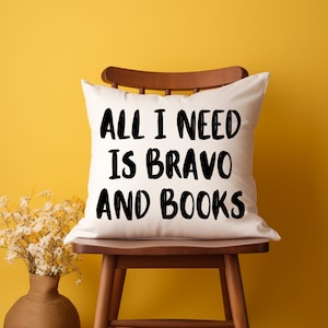 May include: White pillow with black text that reads "All I need is Bravo and Books" on a wooden chair against a yellow wall.