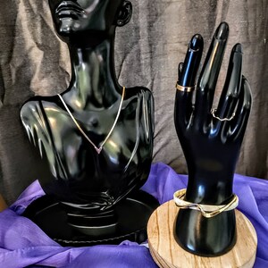 May include: Two black mannequins displaying gold jewelry. A necklace with a V-shaped pendant is on the bust mannequin. A ring and a wavy bangle are on the hand mannequin.