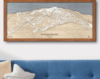 May include: A framed wooden map of Snowmass Mountain in Colorado. The map shows the ski runs and trails on the mountain. The map is hanging on a white wall above a blue sofa.