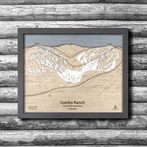 May include: Framed wooden map of Granby Ranch in the Rocky Mountains, Colorado. The map features a detailed layout of ski slopes and trails in a light brown color, with a gray frame. The text "Granby Ranch" and "The Rocky Mountains Colorado" is visible.