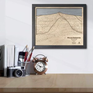 May include: Framed wooden topographic map of Mount Bohemia, with the text "Mount Bohemia The Front Side". The map is in a black frame and is displayed on a light-colored wooden surface. A vintage camera, books, and a copper alarm clock are also visible.