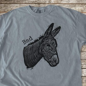 May include: Granite gray t-shirt featuring a black illustration of a donkey's head with the word "Bad" above it. The shirt has rolled-up sleeves and a crew neck. The Buggarnt Print Shop logo is visible in the upper right corner.