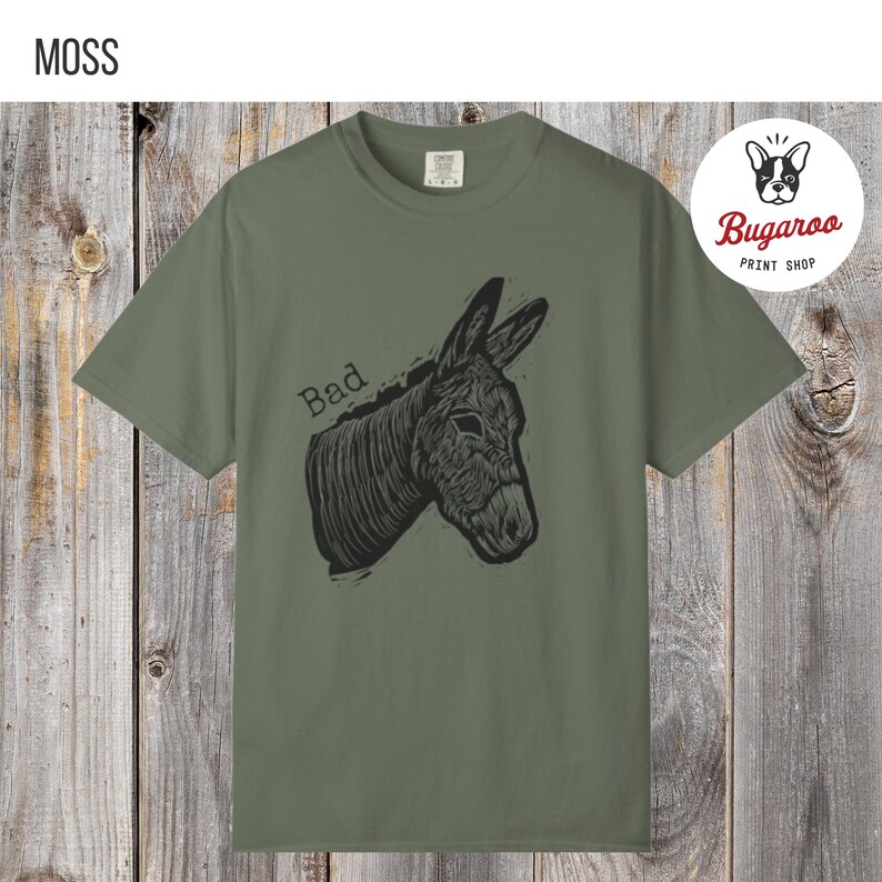 May include: Moss green t-shirt featuring a black illustration of a donkey's head with the word "Bad" above it. The shirt is from the Bugaroo Print Shop.