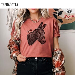 May include: Terracotta-colored t-shirt with a graphic of a donkey head and the word "Bad" above it. The donkey illustration is in dark brown. The shirt is part of a print shop's collection.