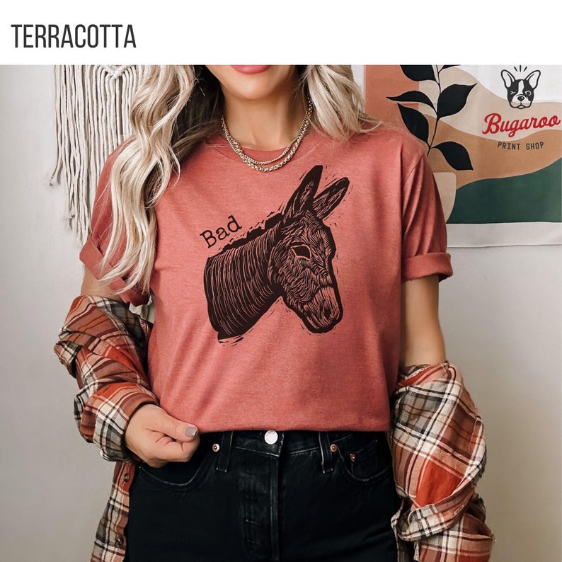 May include: Terracotta-colored t-shirt with a graphic of a donkey head and the word "Bad" above it. The donkey illustration is in dark brown. The shirt is part of a print shop's collection.