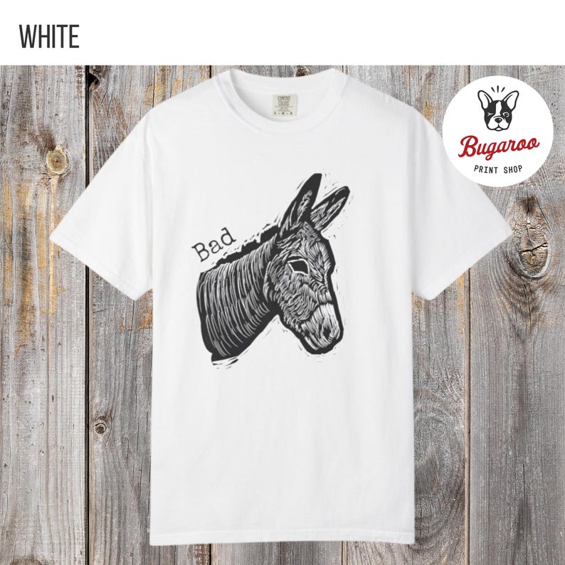 May include: White t-shirt featuring a black and white illustration of a donkey's head with the word "Bad" above it. The shirt has a crew neck and short sleeves. The Bugaroo Print Shop logo is in the upper right corner.