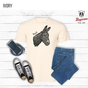 May include: Ivory-colored t-shirt with a black and white illustration of a donkey and the word "Bad". The shirt is paired with blue jeans and black and white sneakers. A smartphone and a charging cable are also visible.