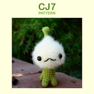 Puede incluir: Un juguete amigurumi de crochet blanco con piernas verdes y una parte superior verde. El juguete tiene ojos negros y una pequeña boca negra.