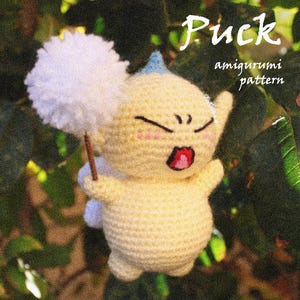 Op de afbeelding: Een gehaakte amigurumi-figuur in lichtgeel, met een blauwe haartooi en roze wangen. De figuur houdt een wit, pluizig voorwerp op een stok vast. De woorden "Puck" en "amigurumi pattern" zijn in het wit geschreven.