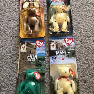 May include: Four Ty Beanie Baby bears in their original packaging. The bears are Britannia, Glory, Erin, and Maple. Each bear is a different color and has a tag attached. The packaging features images of landmarks and flags.