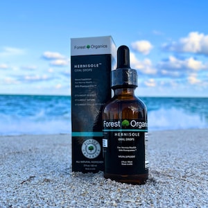 May include: A brown glass bottle with a dropper, labeled "Forest Organics Hernisole Oral Drops" for hernia health with Plaquenil. The bottle is sitting on a sandy beach with a blue ocean and sky in the background. The bottle is next to a box with the same label.