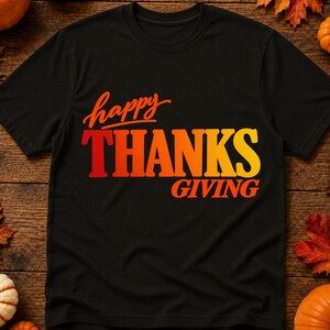 May include: A black short-sleeved t-shirt featuring the text "happy THANKS GIVING" printed in a gradient of orange, red, and yellow. The word "happy" is in a script font, while "THANKS" and "GIVING" are in a bold, sans-serif typeface.