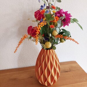 Op de afbeelding: Een decoratieve oranje vaas met een golvend patroon bevat een levendig bloemstuk. Het boeket bevat rode rozen, paarse dahlia's, oranje accenten en groen blad. De vaas staat op een houten oppervlak.