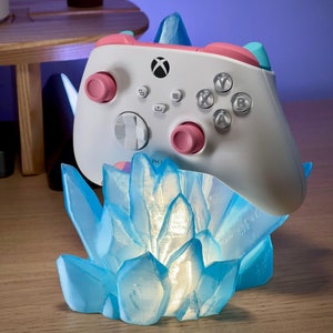 Könnte beinhalten: Ein weißer Xbox-Controller mit rosa und blauen Akzenten wird auf einem blauen kristallförmigen Ständer präsentiert. Der Ständer ist mit einem sanften blauen Licht beleuchtet.