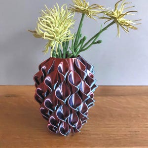Könnte beinhalten: Eine dekorative Vase mit einem einzigartigen, geschichteten Design in Burgunder- und Blautönen. Die Vase enthält mehrere gelbe, stachelige Blumen mit grünen Stielen, die einen auffälligen Kontrast bilden. Die Vase ist etwa 15 cm hoch.