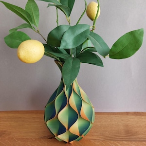 Op de afbeelding: Een decoratieve vaas met een groen en goudkleurig golfpatroon houdt een citroentak met groene bladeren en twee gele citroenen vast. De vaas staat op een houten oppervlak.