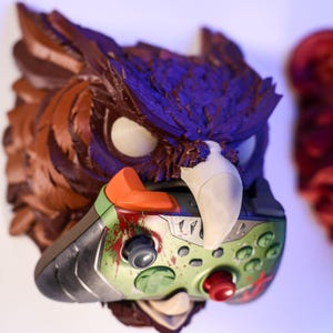 May include: A detailed, hand-painted owl head sculpture in shades of brown, purple, and white, holding a green and silver video game controller with red and orange accents. The controller has a splatter design.