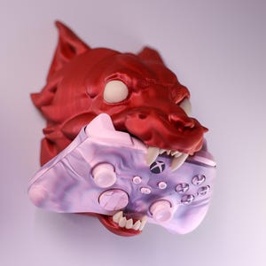 Może przedstawiać: Czerwona głowa wilka zamontowana na ścianie z różowo-fioletowym kontrolerem do gier wideo w paszczy.