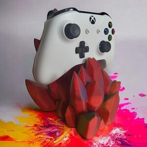 May include: A white Xbox controller sits on a 3D printed red crystal-shaped stand. The stand is on a colorful background with red, yellow, pink, and purple paint splatters.