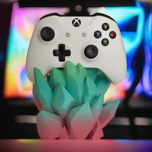 Può includere: Un controller Xbox bianco è posizionato su un supporto a forma di cristallo di colore verde acqua e verde. Il supporto è in plastica e presenta un effetto sfumato.