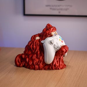 May include: A red, textured monster-like figurine holding a white video game controller. The figurine has sharp teeth and a detailed, fur-like appearance. The controller features colored buttons and directional controls.