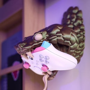 Pode incluir: Um suporte de parede de cabeça de dinossauro impresso em 3D de cor marrom e verde com um controle de videogame branco em sua boca. O controle tem botões rosa e um acento azul e rosa na parte superior.