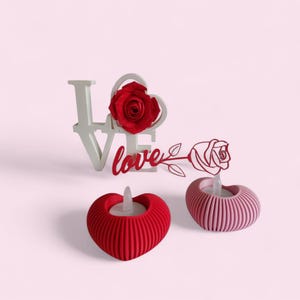 May include: A Valentine's Day-themed arrangement featuring a white "love" sign with a red rose, a red heart-shaped candle holder, and a pink candle holder. The objects are set against a pink background.