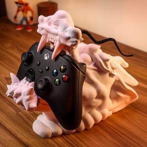 Puede incluir: Una figura de dragón blanca impresa en 3D con un mando de Xbox negro en su boca. El dragón está sentado sobre una superficie de madera.