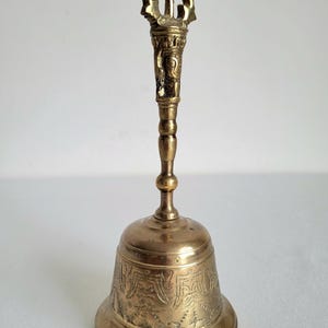 May include: A vintage brass bell with an ornate handle and a detailed, engraved design on the bell's body. The bell has a golden hue and a decorative top. The bell is approximately 6 inches tall.