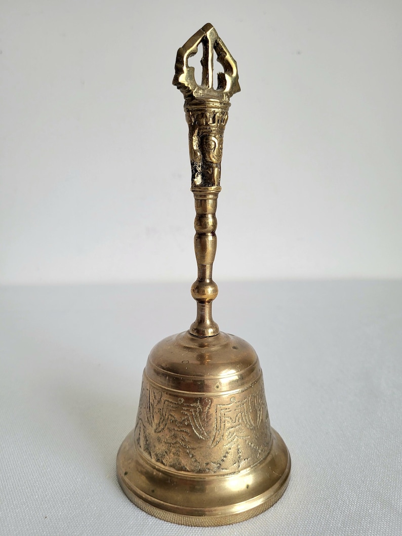 May include: A vintage brass bell with an ornate handle and a detailed, engraved design on the bell's body. The bell has a golden hue and a decorative top. The bell is approximately 6 inches tall.