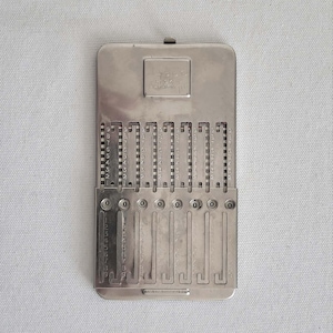 May include: A vintage silver-toned Curta calculator, a mechanical calculator. The device features a rectangular design with multiple number dials and a crank. The top has the word "Curta" engraved. The calculator is designed for mathematical calculations.
