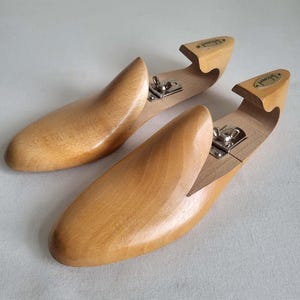May include: Two wooden shoe trees with a natural finish. Each tree has a curved toe and a heel piece with a metal hinge. The heel piece has a small, oval-shaped label. The shoe trees are designed to maintain the shape of shoes.