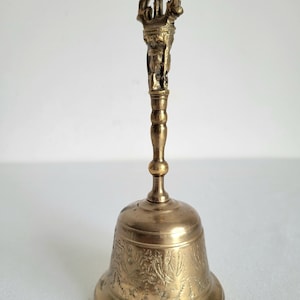 May include: A brass bell with an ornate handle and detailed carvings. The bell has a flared base and a detailed handle with a figure at the top. The bell is a warm gold color and appears to be made of metal.
