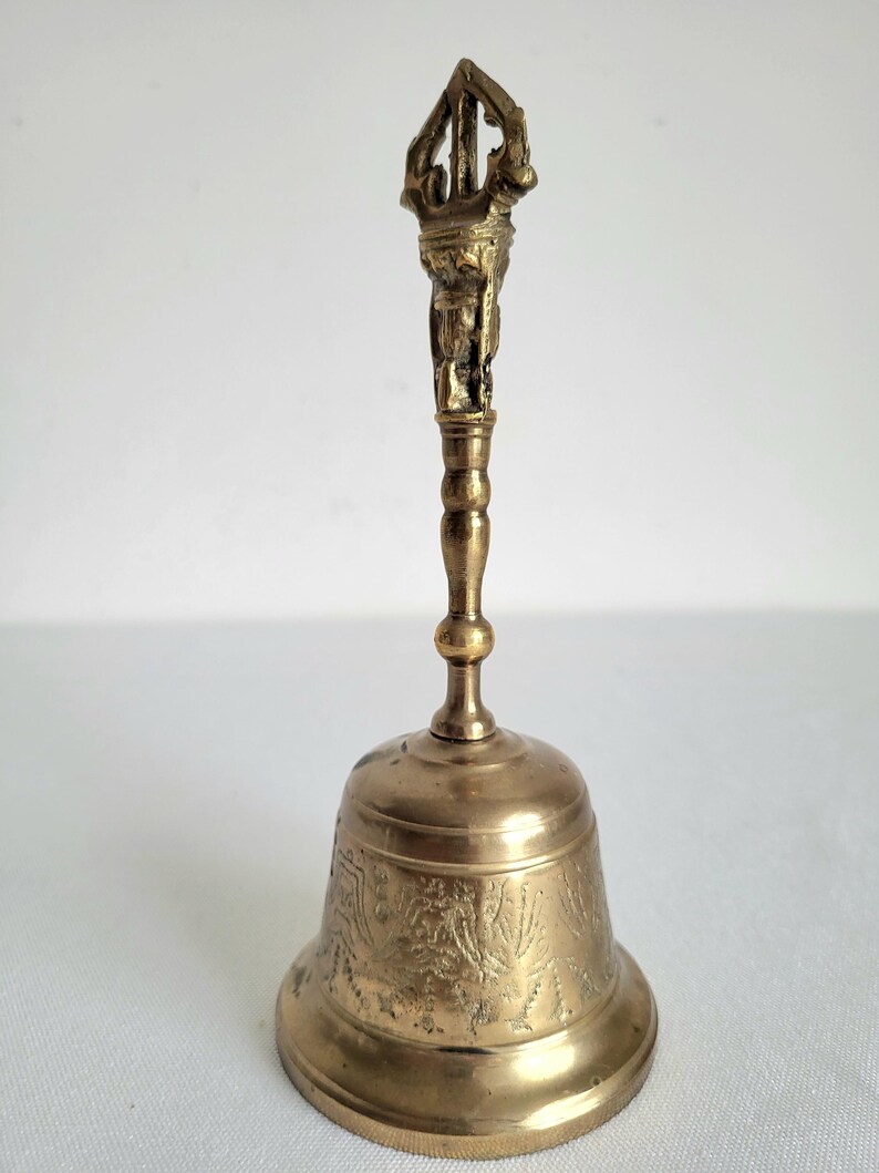 May include: A brass bell with an ornate handle and detailed carvings. The bell has a flared base and a detailed handle with a figure at the top. The bell is a warm gold color and appears to be made of metal.