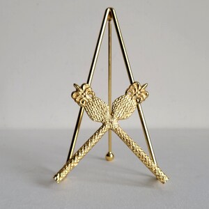 May include: A gold metal display stand with a triangular base and two decorative, crossed arms. The arms are textured and have a stylized design at the ends.