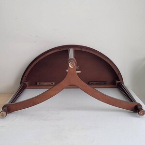 Op de afbeelding: Een halfronde houten tafel met een donkerbruin tafelblad en een lichtbruine, driepotige basis. De tafel heeft een klassiek ontwerp, waarbij de poten naar buiten uitsteken voor stabiliteit. Het tafelblad is ongeveer 76 cm breed.