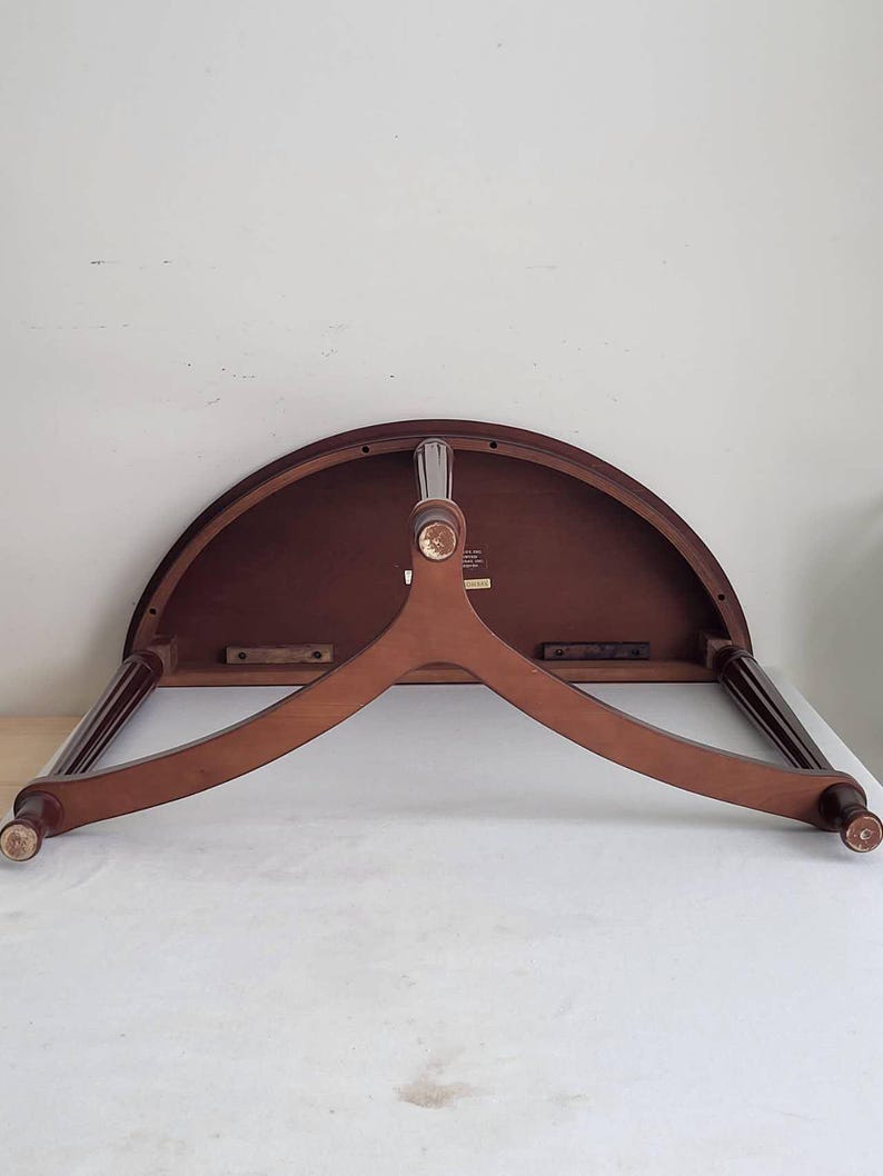 Op de afbeelding: Een halfronde houten tafel met een donkerbruin tafelblad en een lichtbruine, driepotige basis. De tafel heeft een klassiek ontwerp, waarbij de poten naar buiten uitsteken voor stabiliteit. Het tafelblad is ongeveer 76 cm breed.