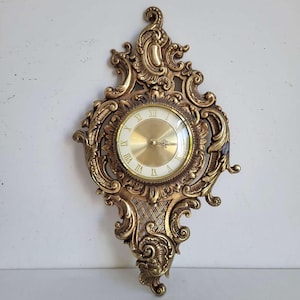 May include: A wall clock featuring an ornate, gold-toned frame with intricate scrollwork and lattice detailing. The clock face is circular with a metallic gold center, a white outer ring, and black Roman numerals. It has two decorative hands.