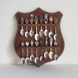 May include: A collection of decorative spoons displayed on a dark brown wooden shield-shaped rack. The spoons have various designs and colors, with silver and gold tones. The rack is mounted on a white wall.