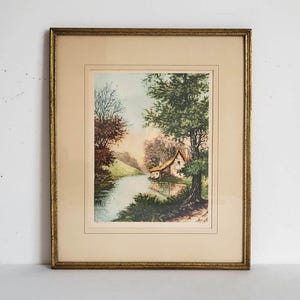 May include: A framed landscape print featuring a small cottage situated beside a river, surrounded by green trees and foliage. The artwork is enclosed in a rectangular gold-toned frame with a cream-coloured mat border, showcasing a serene scene.