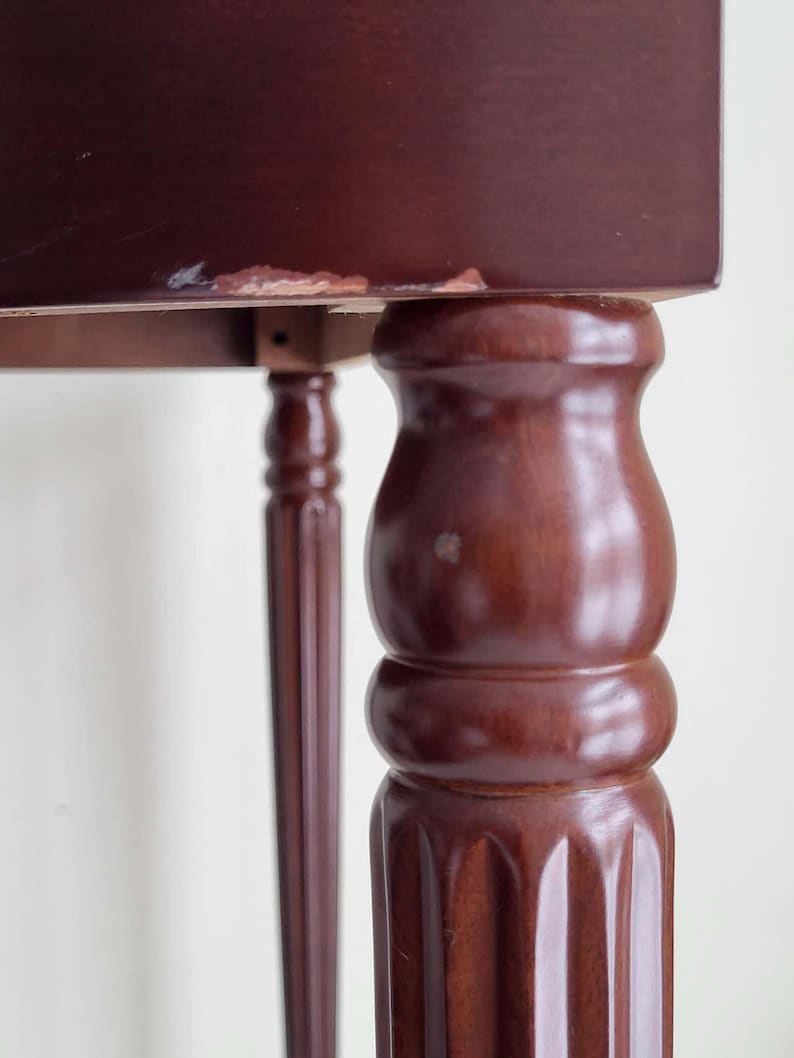 Op de afbeelding: Close-up van een donkerbruine houten tafel. De tafel heeft een rechthoekig blad en sierlijke, gedraaide poten. E&eacute;n poot is een dikke, afgeronde kolom, terwijl een andere een slank, gefluteerd ontwerp heeft. De afwerking van de tafel heeft een rijke, warme tint.