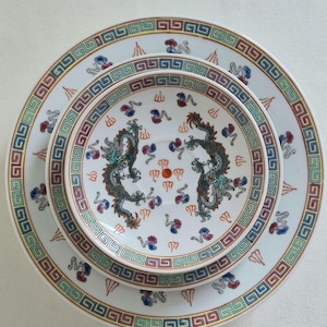 May include: A white ceramic plate and saucer set featuring a colorful dragon motif in green, red, and blue. Both pieces are decorated with a geometric Greek key border in gold, blue, and red, and small scattered cloud-like symbols in red and blue.