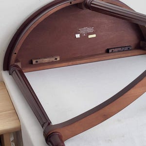 Op de afbeelding: Een donker kersenkleurig, halfrond tafeltje met twee poten. De onderkant van de tafel is zichtbaar, met de houtnerf en constructiedetails. De poten zijn donkerder kersenkleurig en hebben een gedraaid ontwerp.