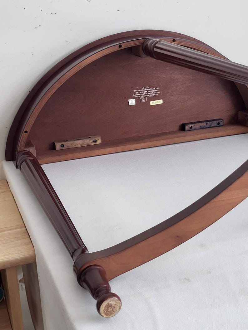 Op de afbeelding: Een donker kersenkleurig, halfrond tafeltje met twee poten. De onderkant van de tafel is zichtbaar, met de houtnerf en constructiedetails. De poten zijn donkerder kersenkleurig en hebben een gedraaid ontwerp.