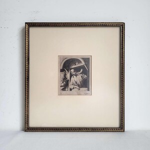 May include: A framed black and white etching featuring a person wearing a cap and blowing into a horn. The artwork is centered on a cream-colored mat within a dark, textured frame with a scalloped edge. A signature is visible in the bottom right.