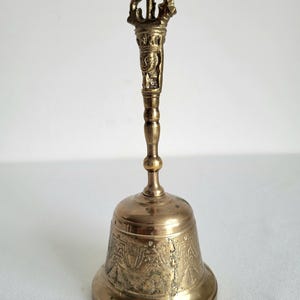 May include: A brass bell with an ornate handle and a detailed, patterned bell. The handle features intricate carvings and a decorative top. The bell is a warm gold color and appears to be made of metal.