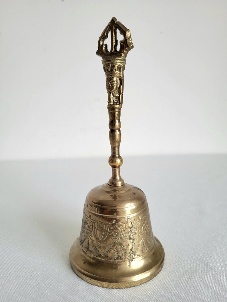 May include: A brass bell with an ornate handle and a detailed, patterned bell. The handle features intricate carvings and a decorative top. The bell is a warm gold color and appears to be made of metal.