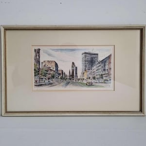 May include: Framed watercolour print of a city street scene. The artwork depicts buildings, cars, and people. The print is in a cream-coloured mount with a gold-coloured frame.